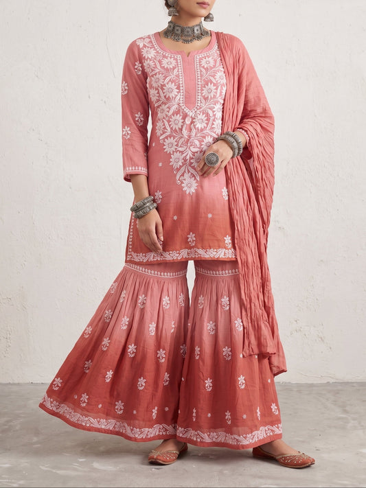 A woman models a three-piece salwar kameez suit in a dusty rose pink color with white floral embroidery. The set includes a long-sleeve tunic, matching wide flared pants, and a draped dupatta, styled with traditional jewelry against a textured off-white wall.