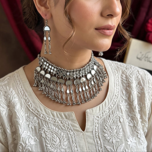 A woman wearing a detailed silver-toned traditional Indian choker necklace with dangling mirrors and a matching earring, styled with an embroidered cream kurta against a background of dark red drapery and flowers.