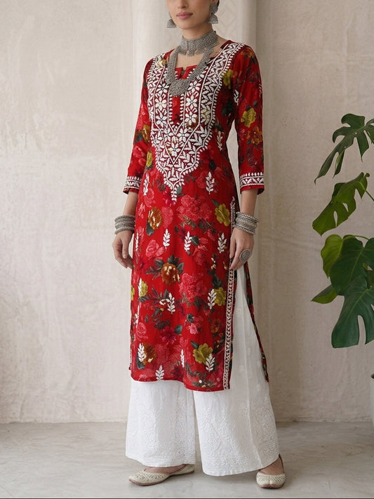 A woman modeling a red floral printed kurta with intricate white embroidery on the neckline, paired with white wide-leg pants, standing in front of a textured beige wall next to a potted green plant.