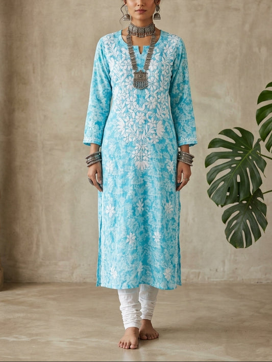 A woman stands barefoot indoors wearing a light blue and white printed kurta with white pants, layered silver jewelry, and a long necklace. The setting has a textured beige wall, a carved wooden pillar on the left, and a green monstera plant on the right.