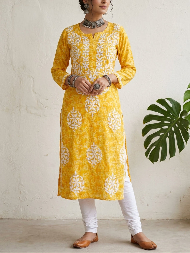 A woman modeling a yellow kurta with white floral embroidery, paired with white pants and brown shoes, standing against a textured off-white wall with a green plant leaf on the right.