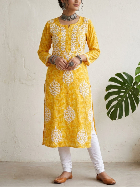 A woman modeling a yellow kurta with white floral embroidery, paired with white pants and brown shoes, standing against a textured off-white wall with a green plant leaf on the right.