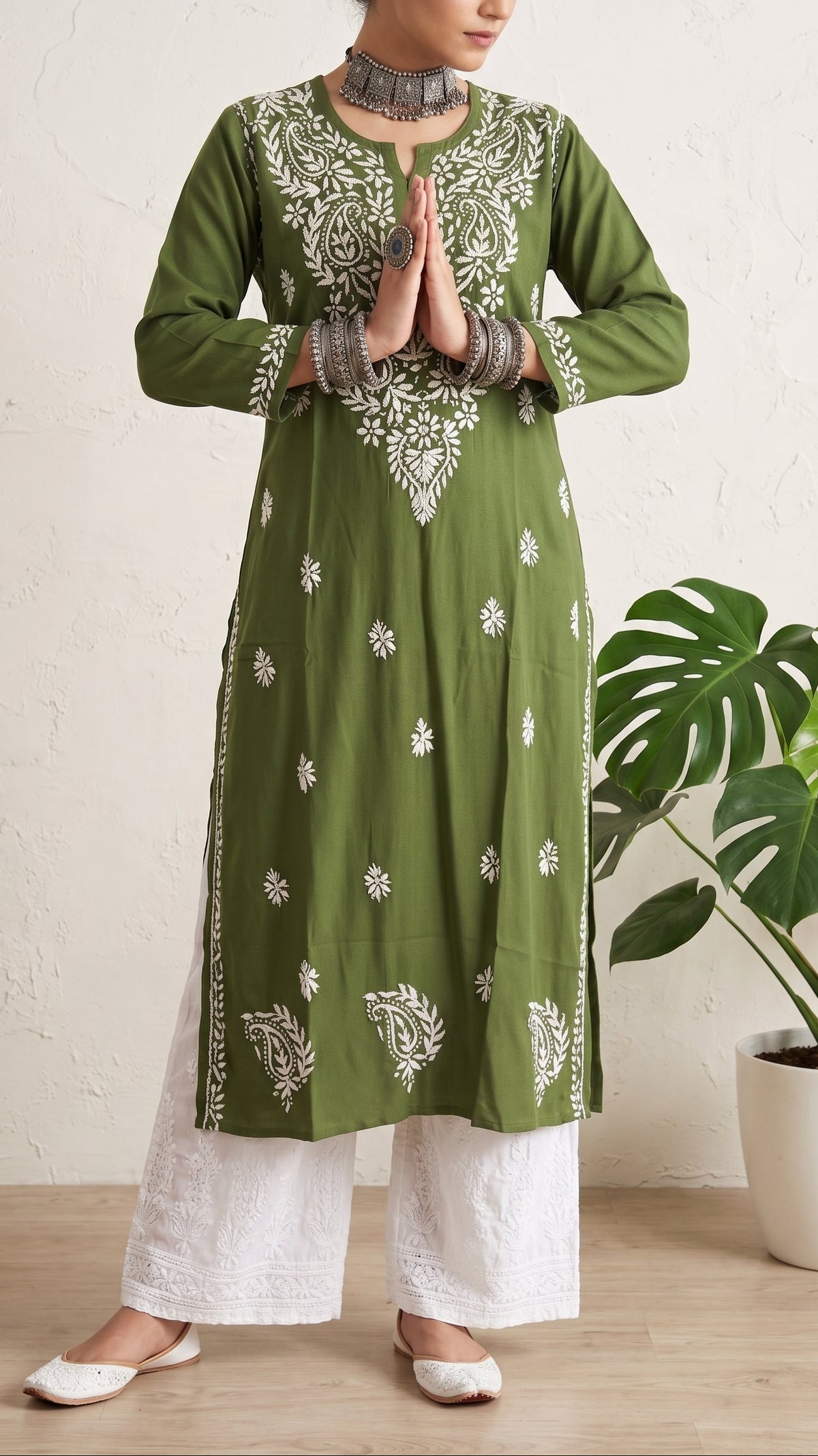 A woman wearing an olive green long kurta with white embroidery, paired with white wide-leg pants and white shoes, standing in a room with a potted plant.