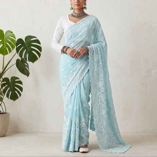 A woman models a light blue saree with white floral embroidery, paired with a white blouse, standing in front of a textured off-white wall next to a potted green plant.