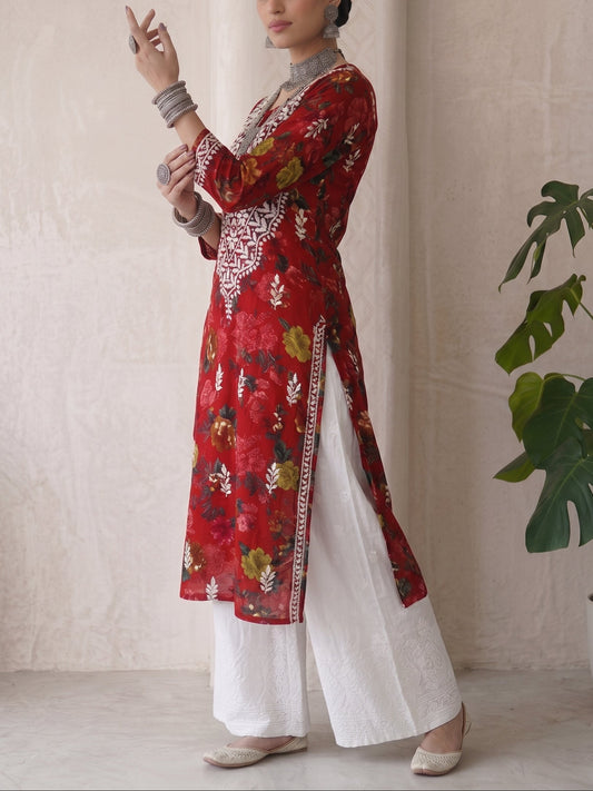 A woman models a red floral printed kurta with white wide-leg pants, standing in a neutral indoor setting with a potted plant in the background.