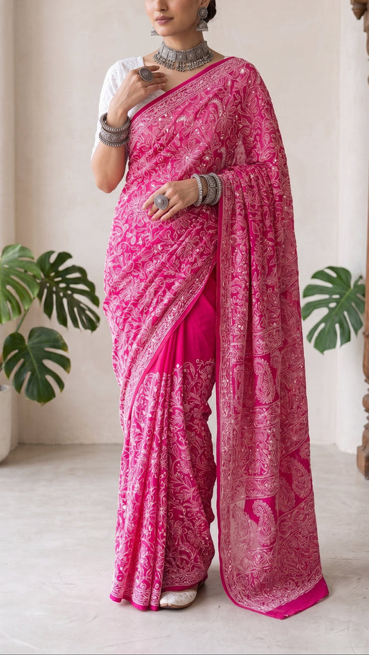 A woman models a bright pink saree with intricate silver embroidery and a matching pink dupatta, paired with a white blouse, in an indoor setting with plants and a wooden pillar.