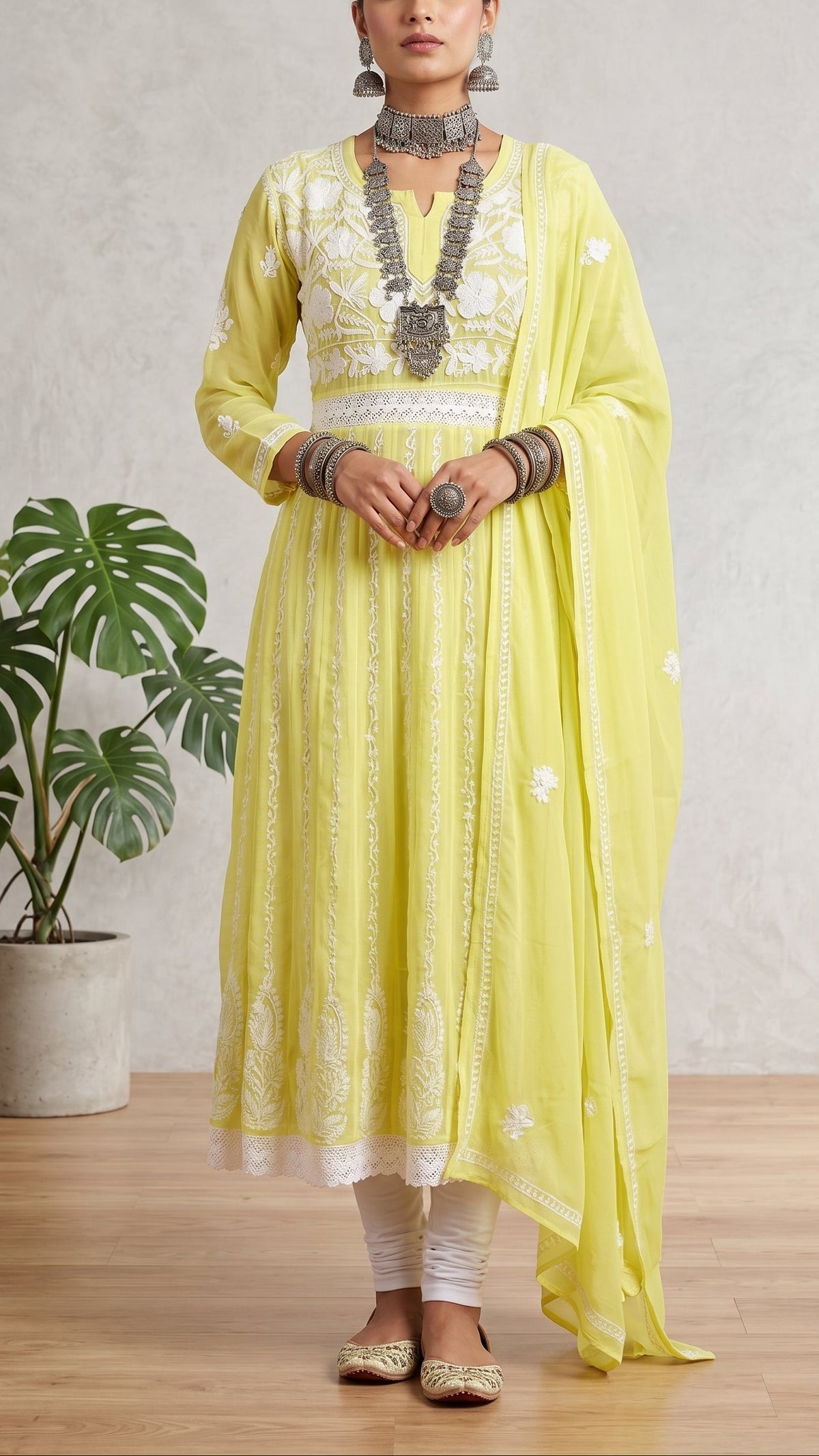 A woman models a light yellow Anarkali suit with white embroidery, white pants, and a matching sheer yellow dupatta, standing in a room with a potted plant and textured wall.