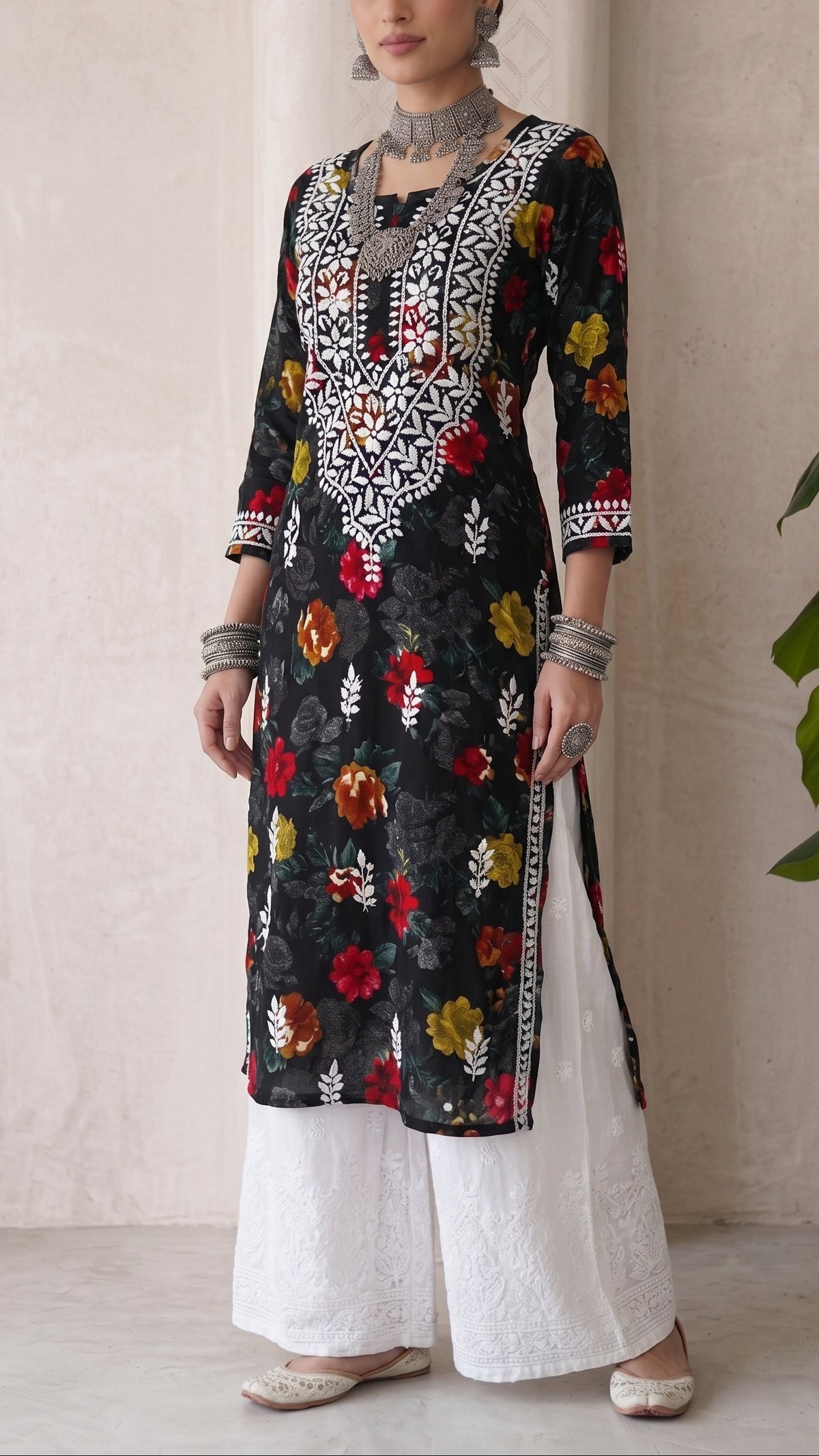 A woman modeling a black embroidered kurta with colorful floral motifs and white wide-leg pants, standing against a neutral beige wall with a green plant leaf visible on the right.