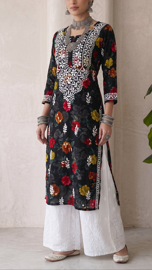 A woman modeling a black embroidered kurta with colorful floral motifs and white wide-leg pants, standing against a neutral beige wall with a green plant leaf visible on the right.