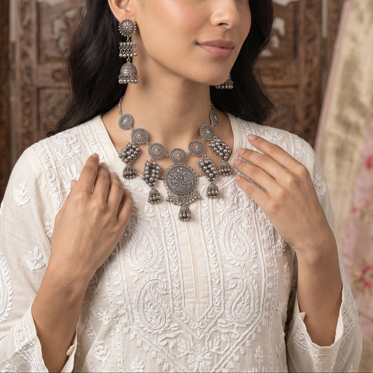 A woman wearing an ornate silver-toned traditional necklace and matching earrings, styled with a white embroidered kurta, set against a background of carved wooden furniture and a pink patterned fabric.