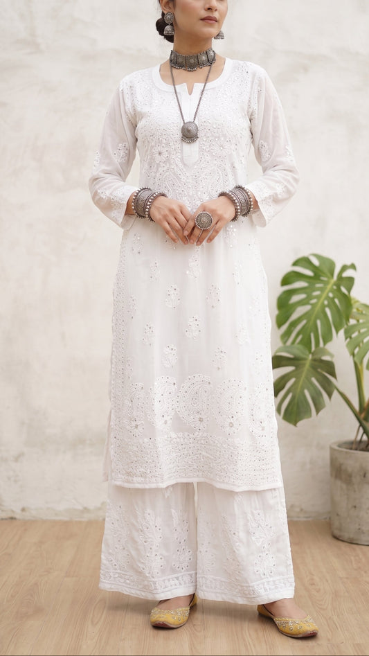A woman modeling a white embroidered kurta and wide-leg pants set with silver jewelry, standing in front of a textured light gray wall next to a potted green plant.